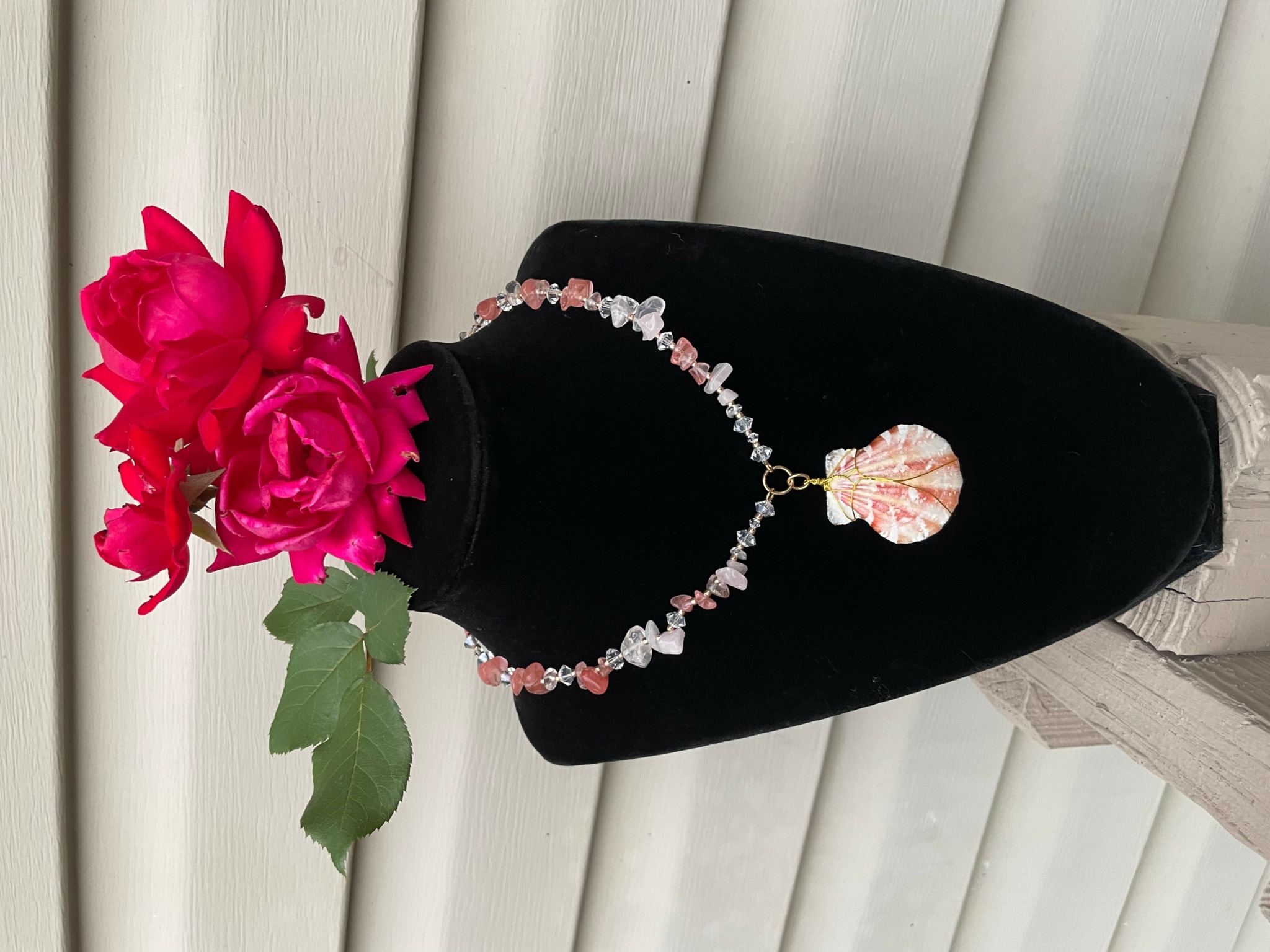 Black mannequin bust with pink flowers as head, wearing a beaded necklace with a large pendant, against a vertical blinds background.