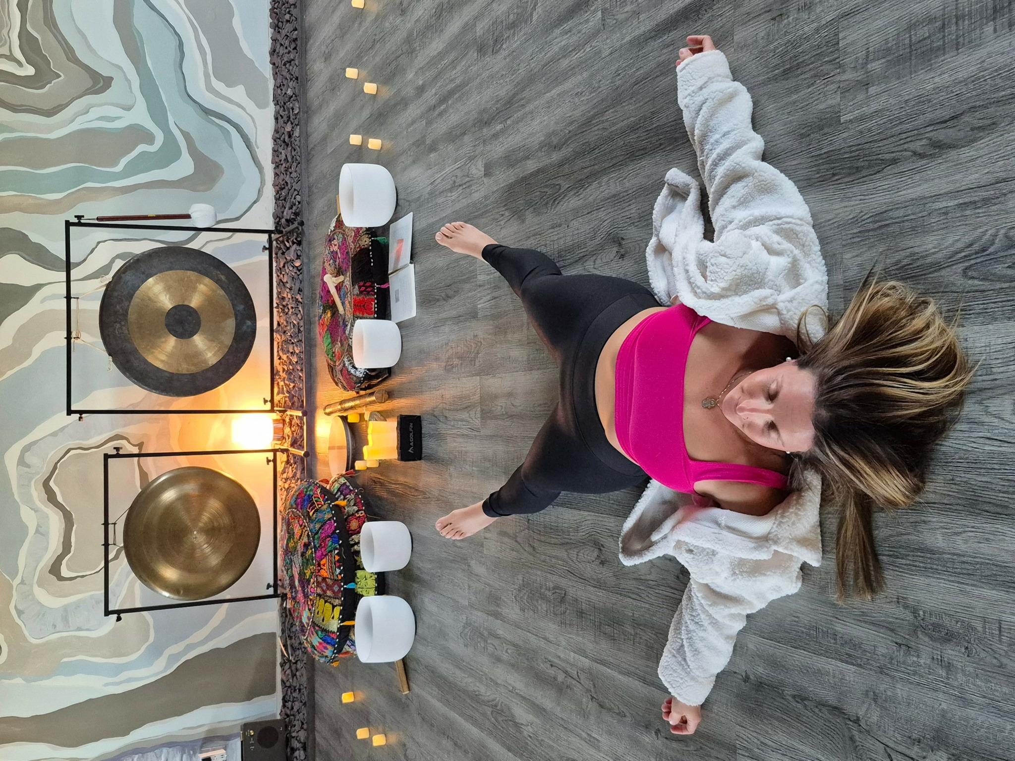 Woman sitting on floor with colorful decorations, a gong, and candles, looking up at the camera, with a gray wooden floor.