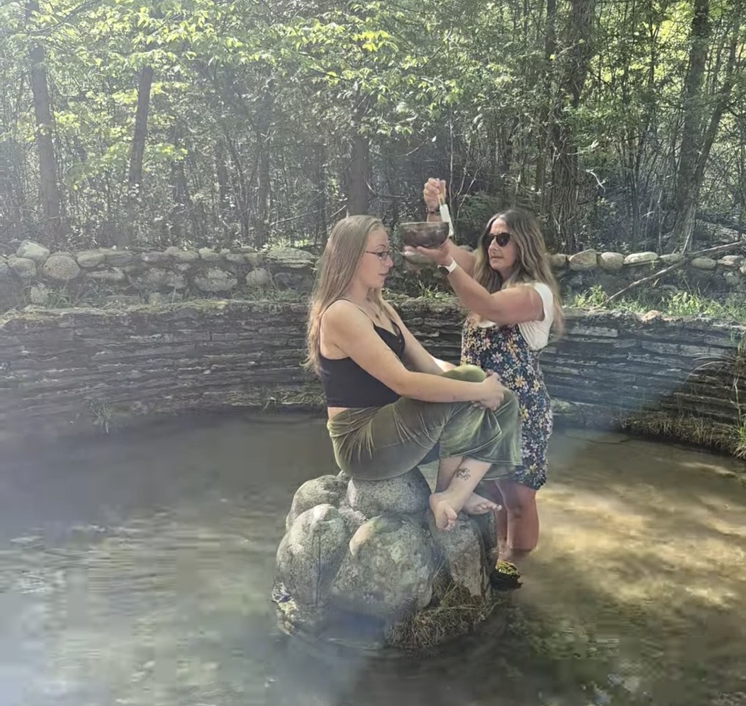 Two women are outdoors near a small pond, one sitting on rocks and the other pouring water over her head, surrounded by trees.