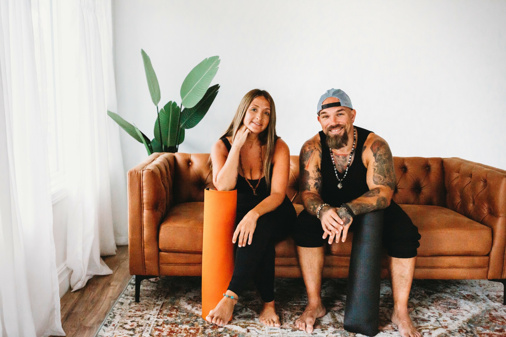 Two people sitting on a brown sofa in a bright room with a large plant, smiling at the camera.