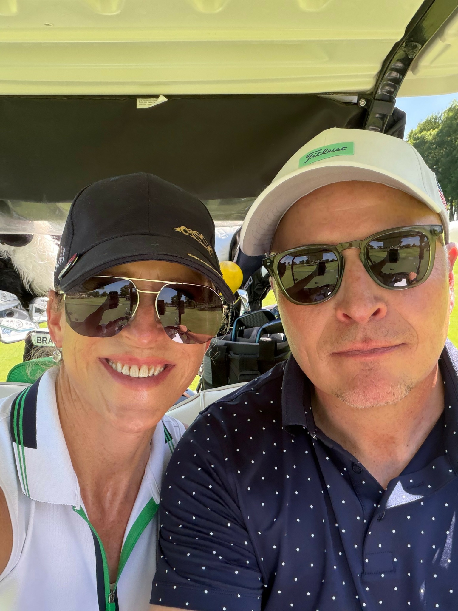 Two people wearing sunglasses and hats, smiling and taking a selfie outdoors with a golf cart in the background.