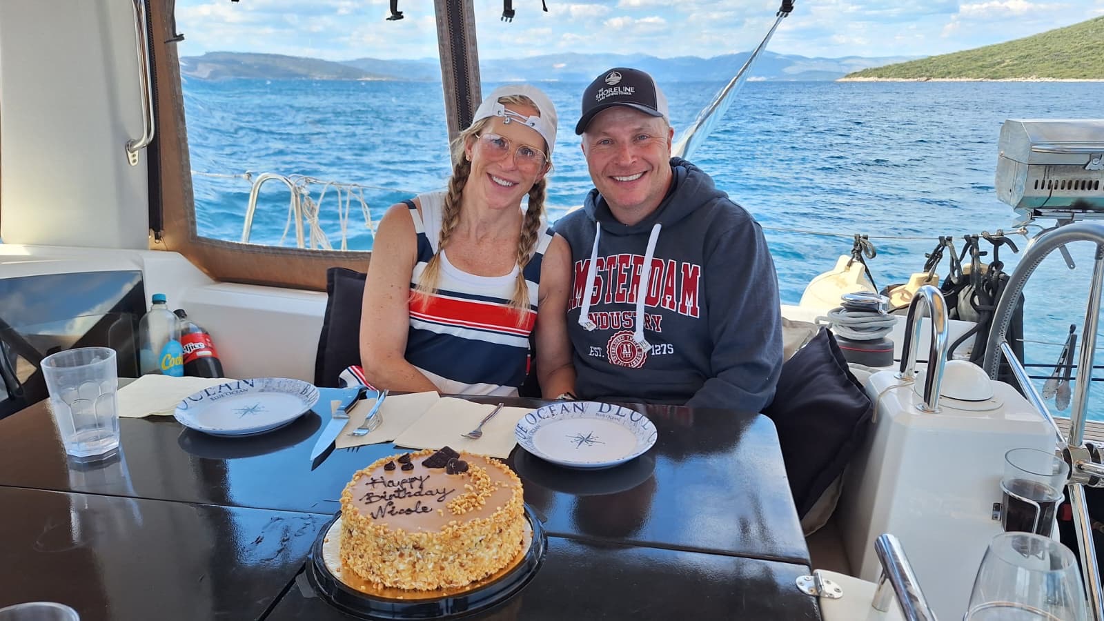 Two people sit at a table on a boat with a cake, smiling, with water and land visible outside the window.