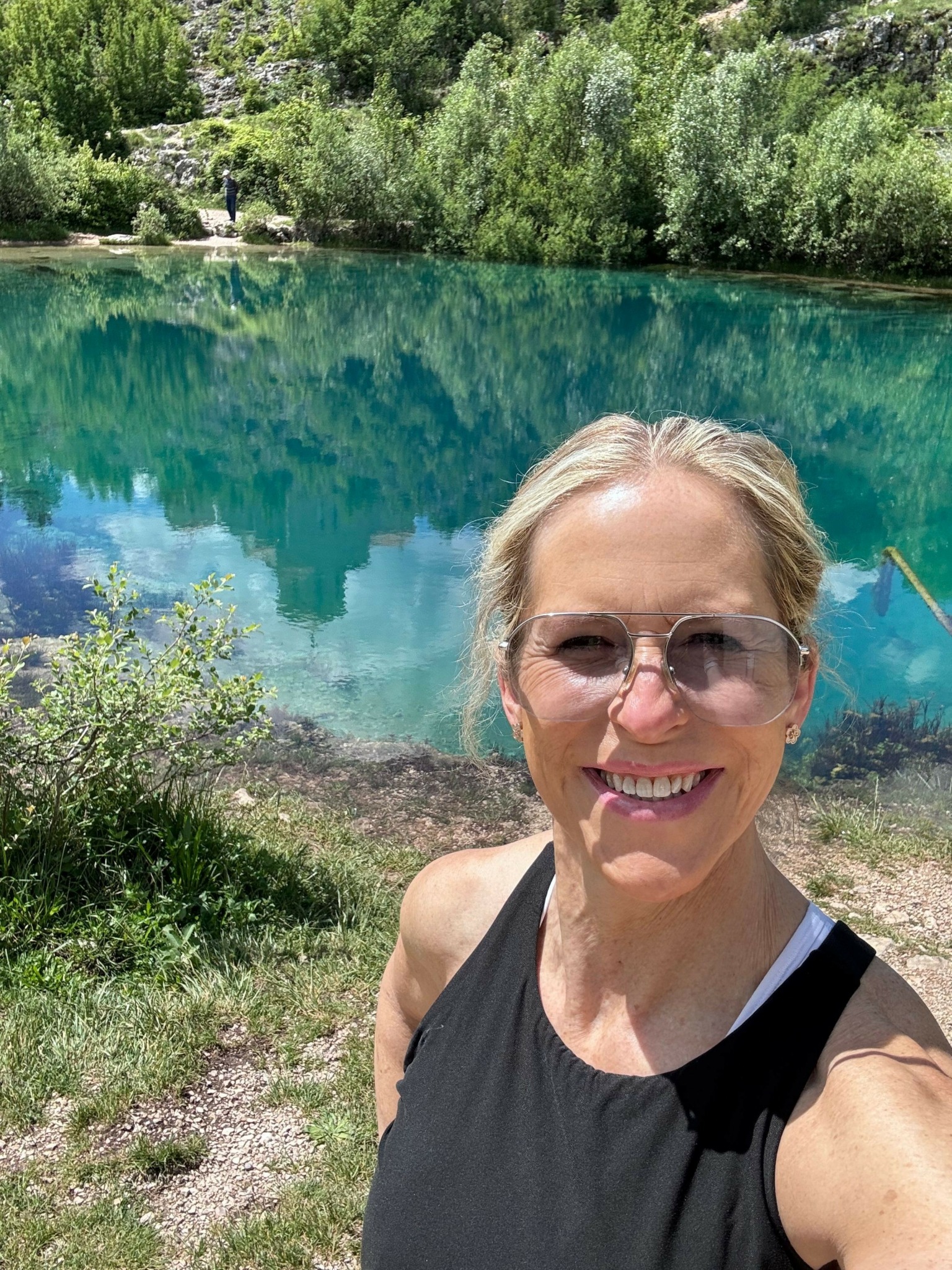 Woman with sunglasses smiling outdoors near a body of water with trees and a city skyline in the background.