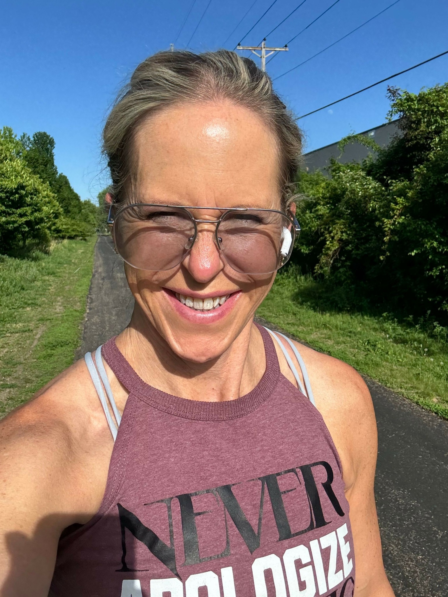 Smiling woman with glasses outdoors, wearing a sleeveless top, with trees and blue sky in background.