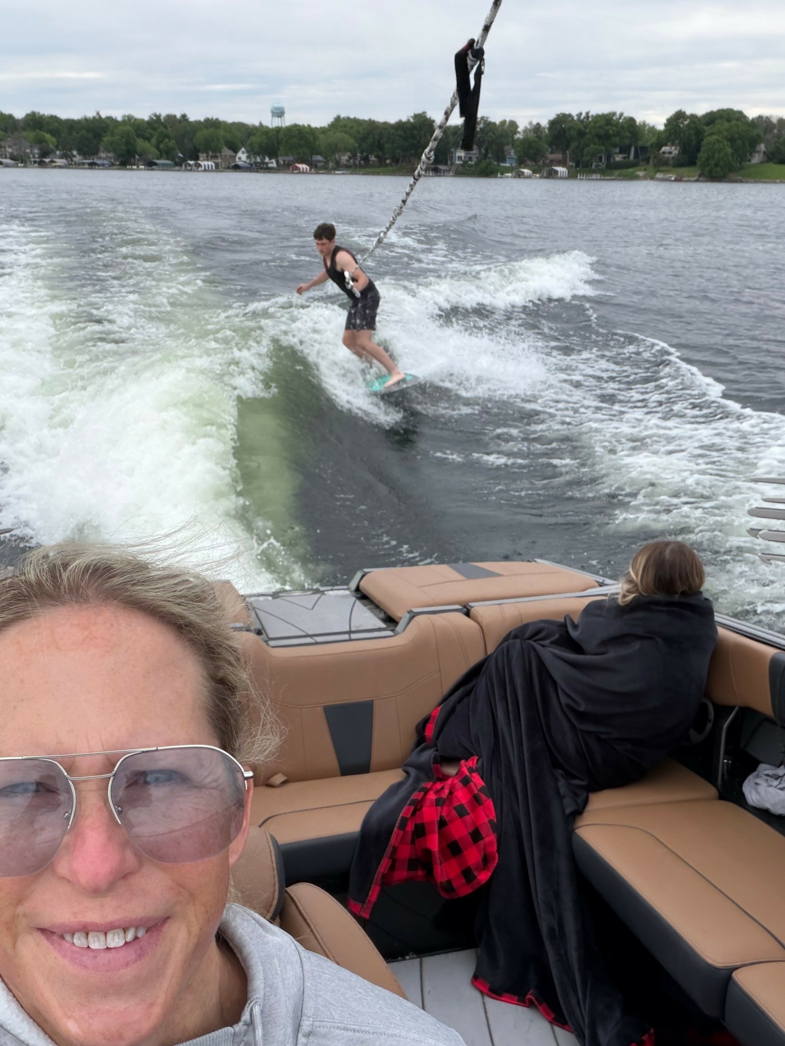 Person wakeboarding on water, boat with two people in the foreground, trees and sky in background.