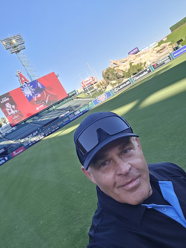 Man wearing a cap and sunglasses taking a selfie at a sports stadium with a green field and large screens in the background.