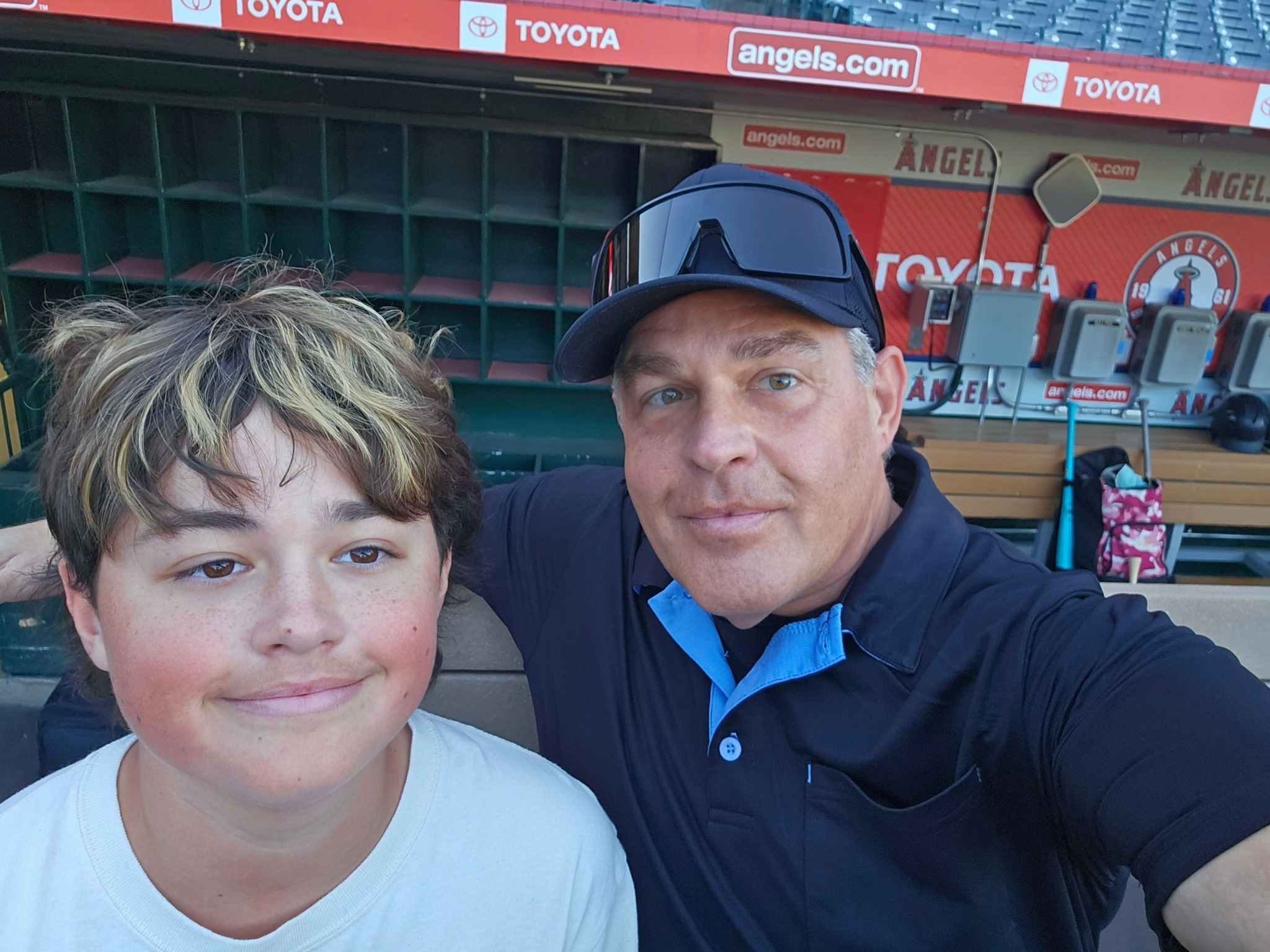 Two people taking a selfie outdoors, one wearing a cap and the other with short hair, smiling at the camera.