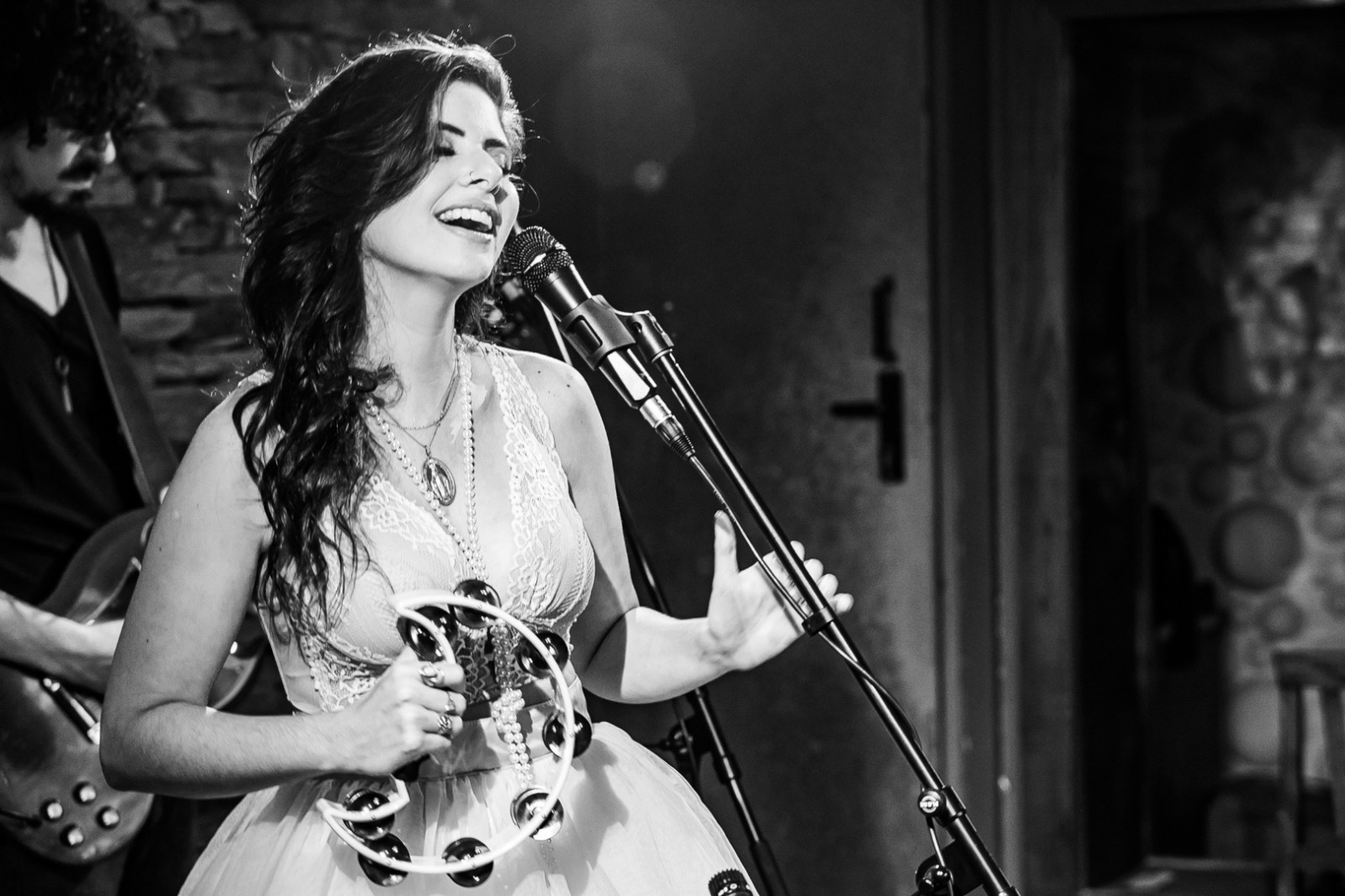 Woman singing into a microphone, holding a tambourine, with a guitarist in the background, in a black and white setting.