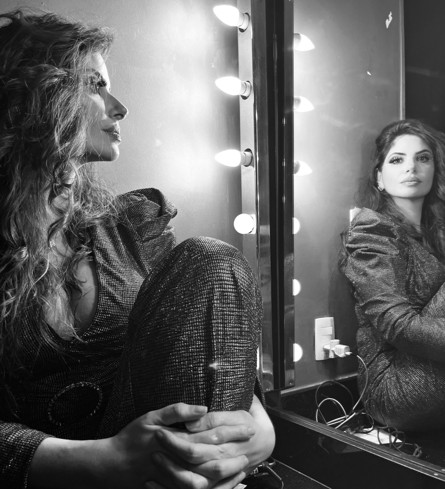 Two women sit in front of a mirror with lights, one looking at her reflection, the other gazing away, both in dark clothing.