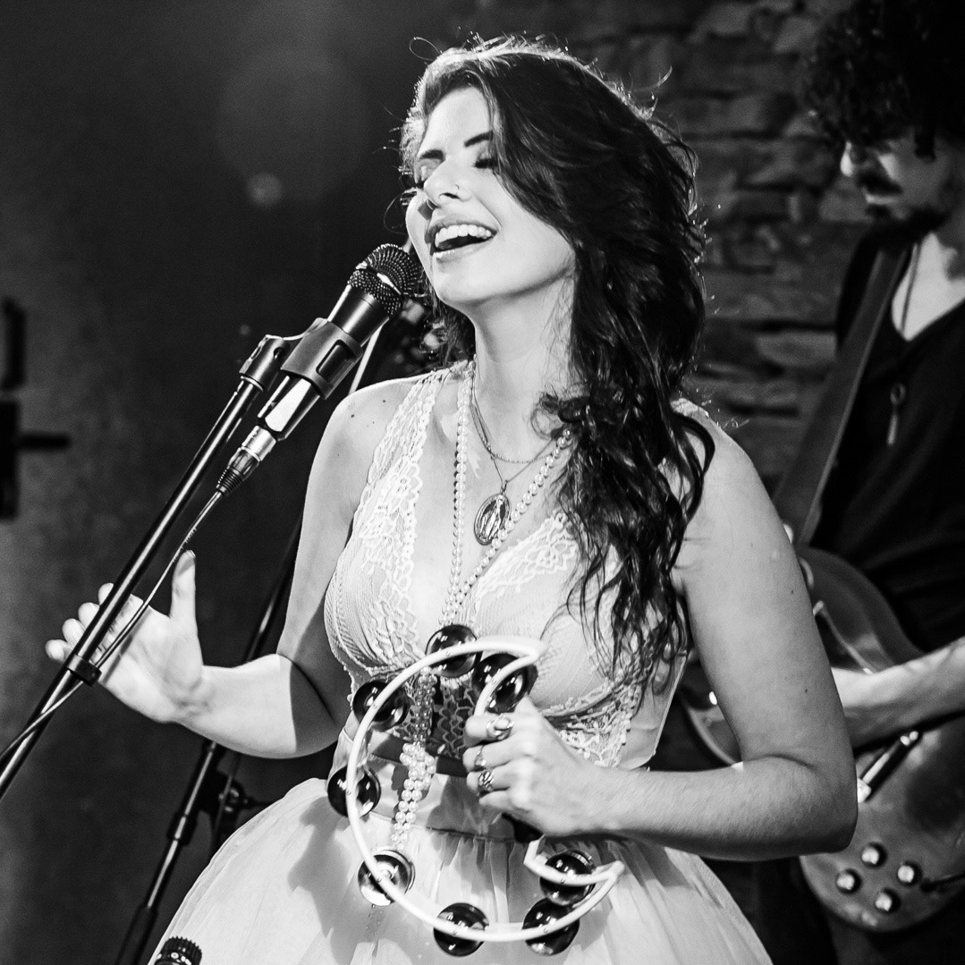 Woman singing with a tambourine in front of a microphone, smiling, with a musician playing guitar in the background.