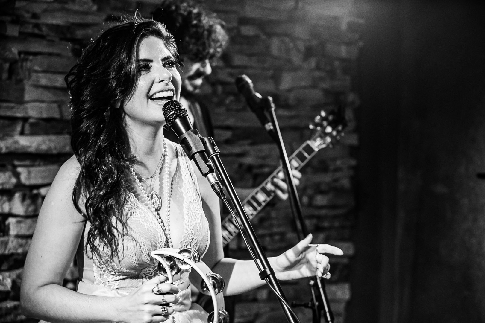 Woman singing into a microphone on stage with a guitarist in the background, brick wall behind her.