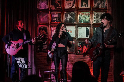 Three musicians perform on stage with framed pictures behind them, one singing and two playing guitars.
