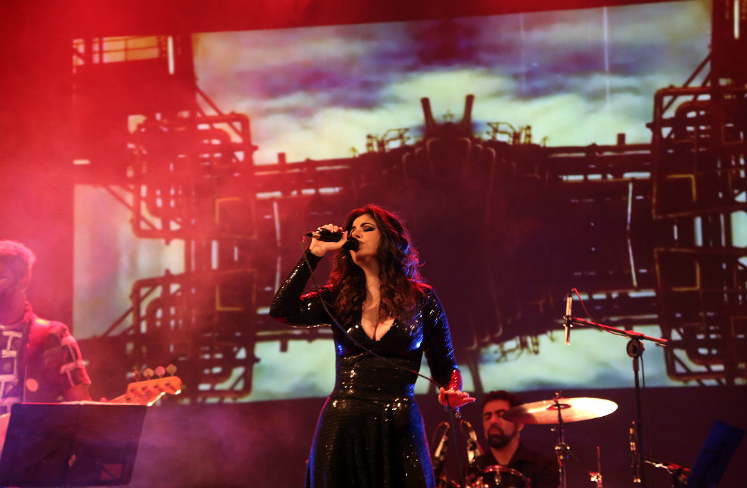 Female singer with long dark hair performs on stage with band, industrial background projection, colorful lighting.