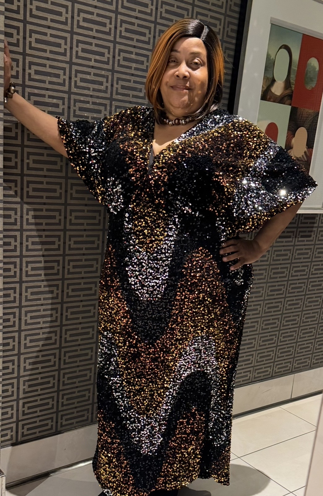 Woman in a black and gold sequined dress standing with one arm extended, smiling, in front of patterned wall and framed artwork.