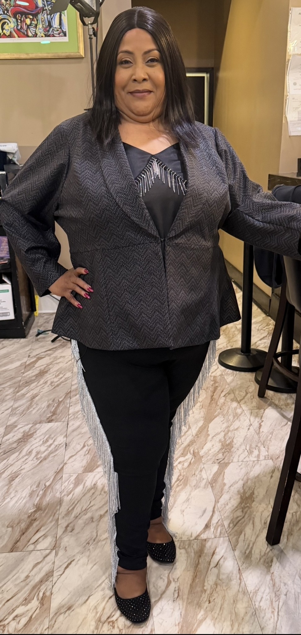Woman standing indoors, wearing a dark blazer, black pants, and heels, with one hand on her hip.