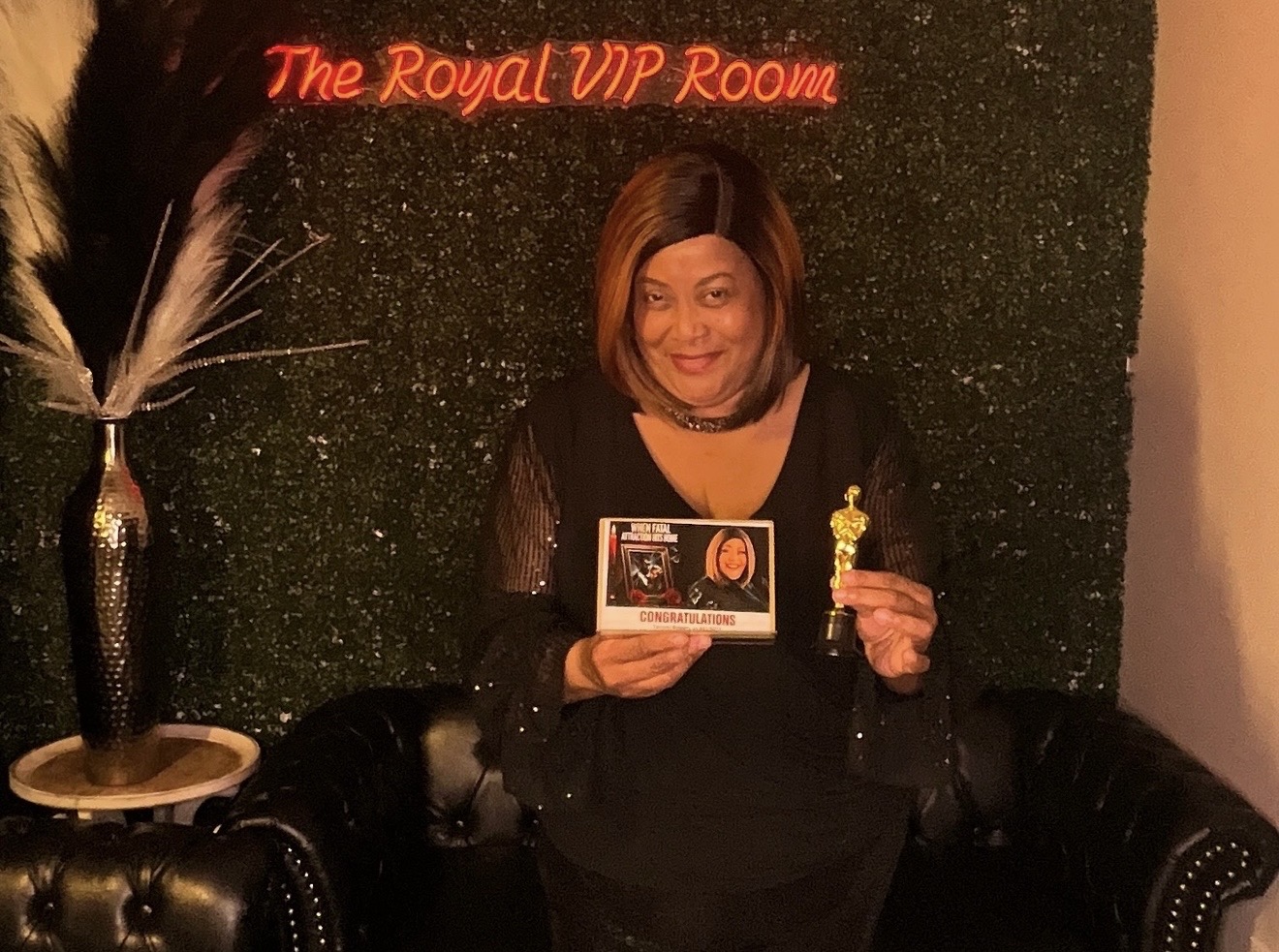 Woman holding a trophy and a certificate in front of a green wall with red text, seated on a black leather chair.