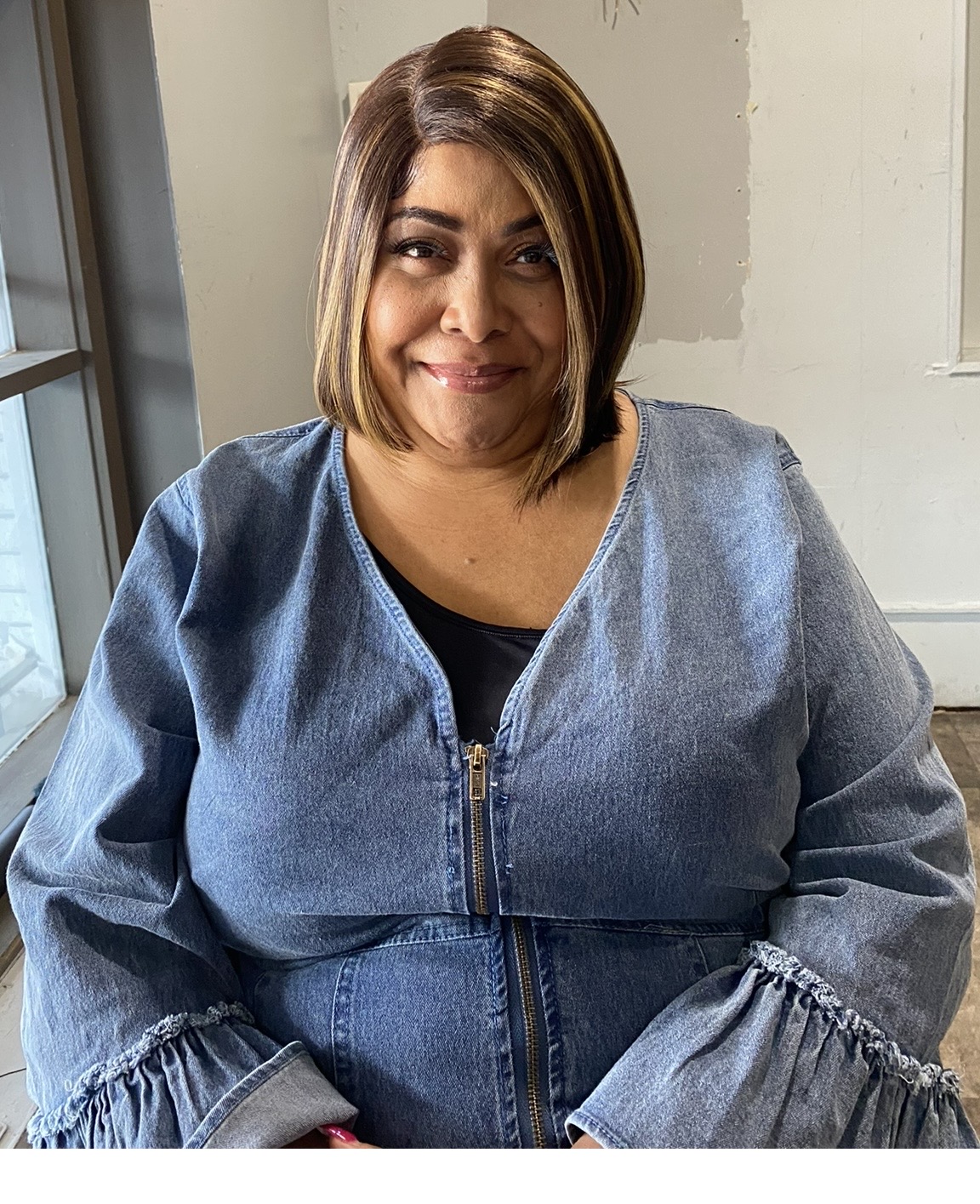 Woman with shoulder-length hair smiling, wearing a denim jacket, sitting indoors near a window.