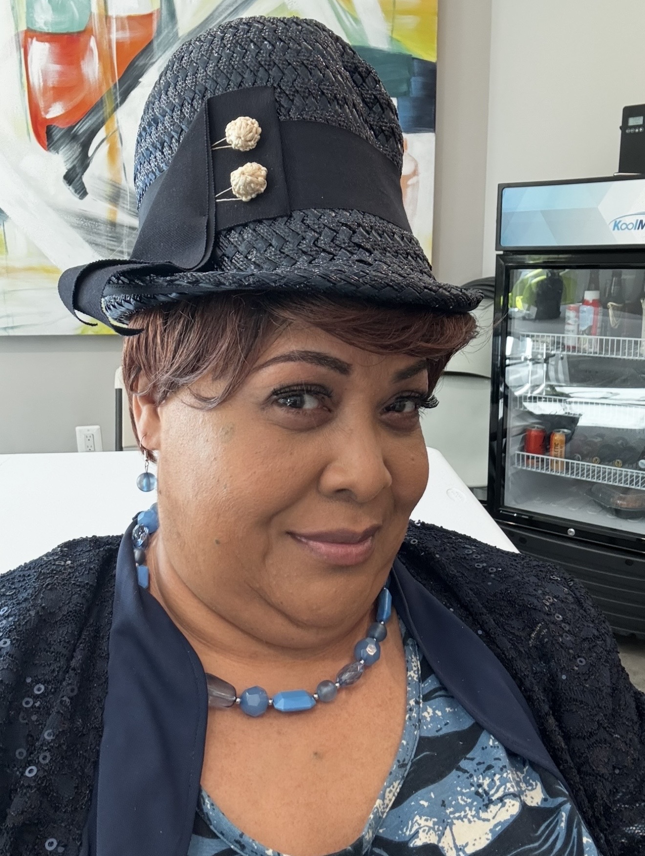 Woman wearing a hat with decorative pins, smiling, indoors with a refrigerator in the background.