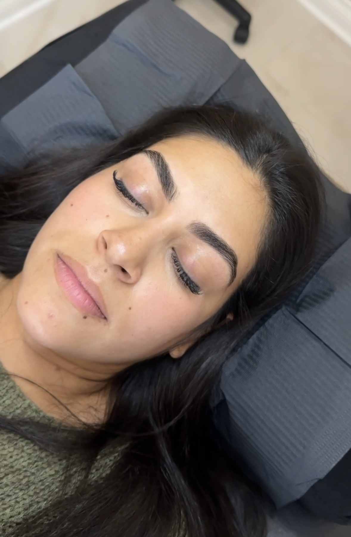 Woman with closed eyes lying on a pillow, with makeup and dark hair, in a relaxed setting.