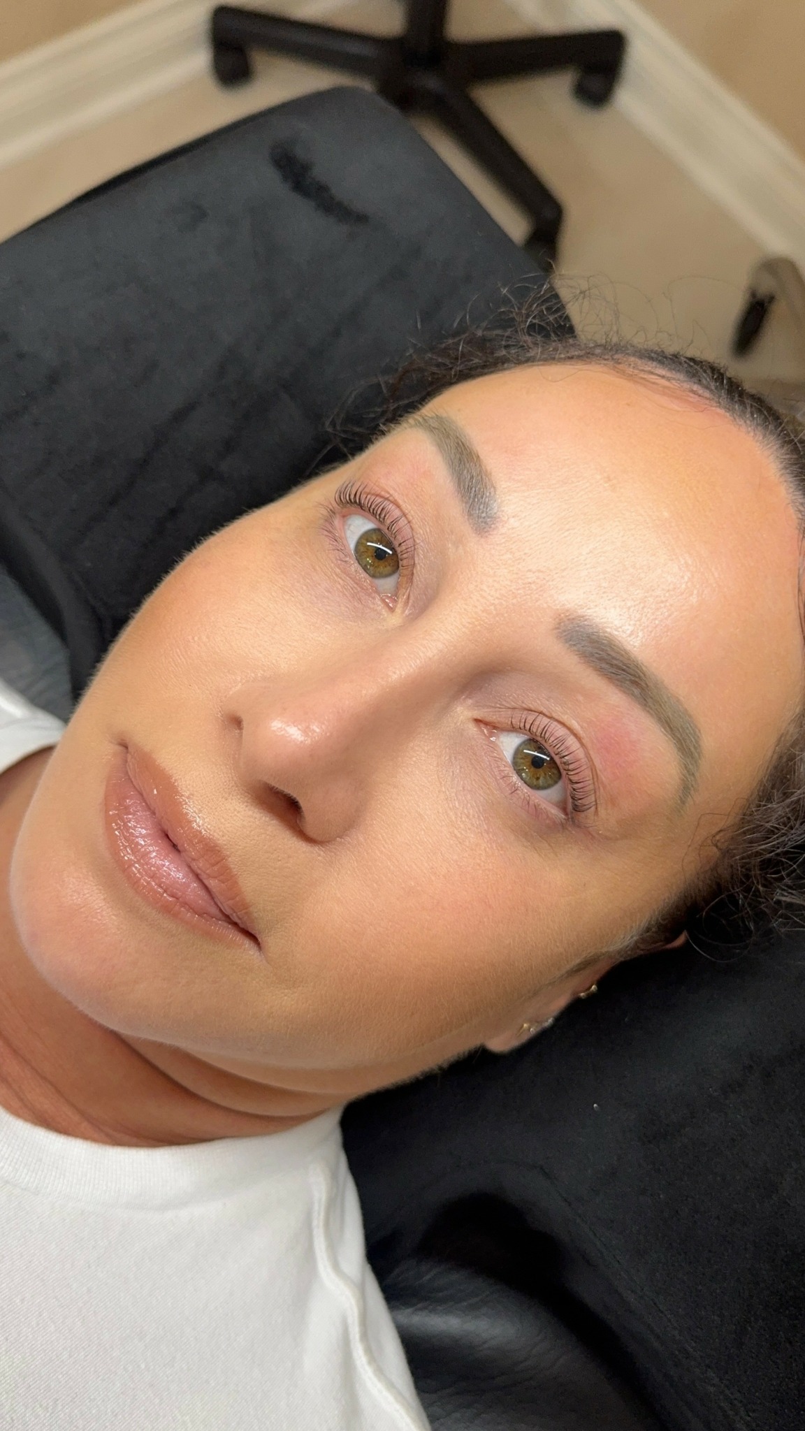 Close-up of a woman's face with makeup, lying down on a black surface, background shows a chair and beige wall.