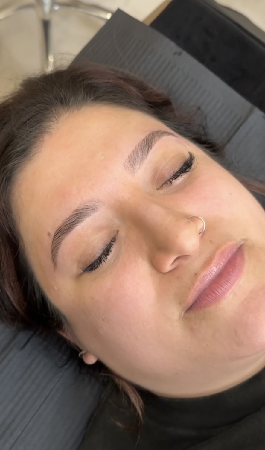 Close-up of a woman with closed eyes, lying down, with a nose piercing and makeup, in a relaxed state.