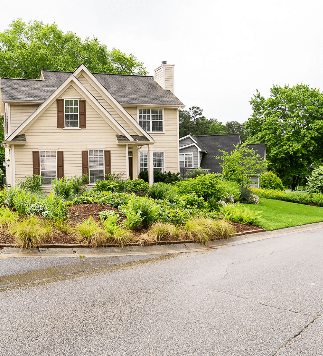 Front yard with garden and house, trees in background, cloudy sky, curb and street in foreground.