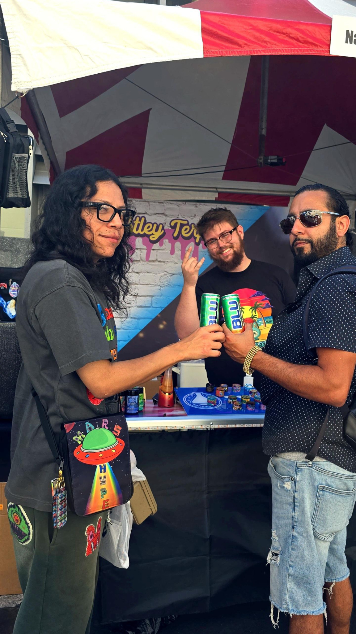 Three people standing outdoors under a tent, holding drinks, with a table and colorful decorations in the background.