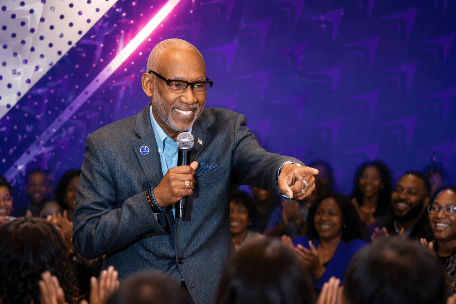 Man in suit speaking into microphone, audience smiling and clapping, purple background, lively event setting.