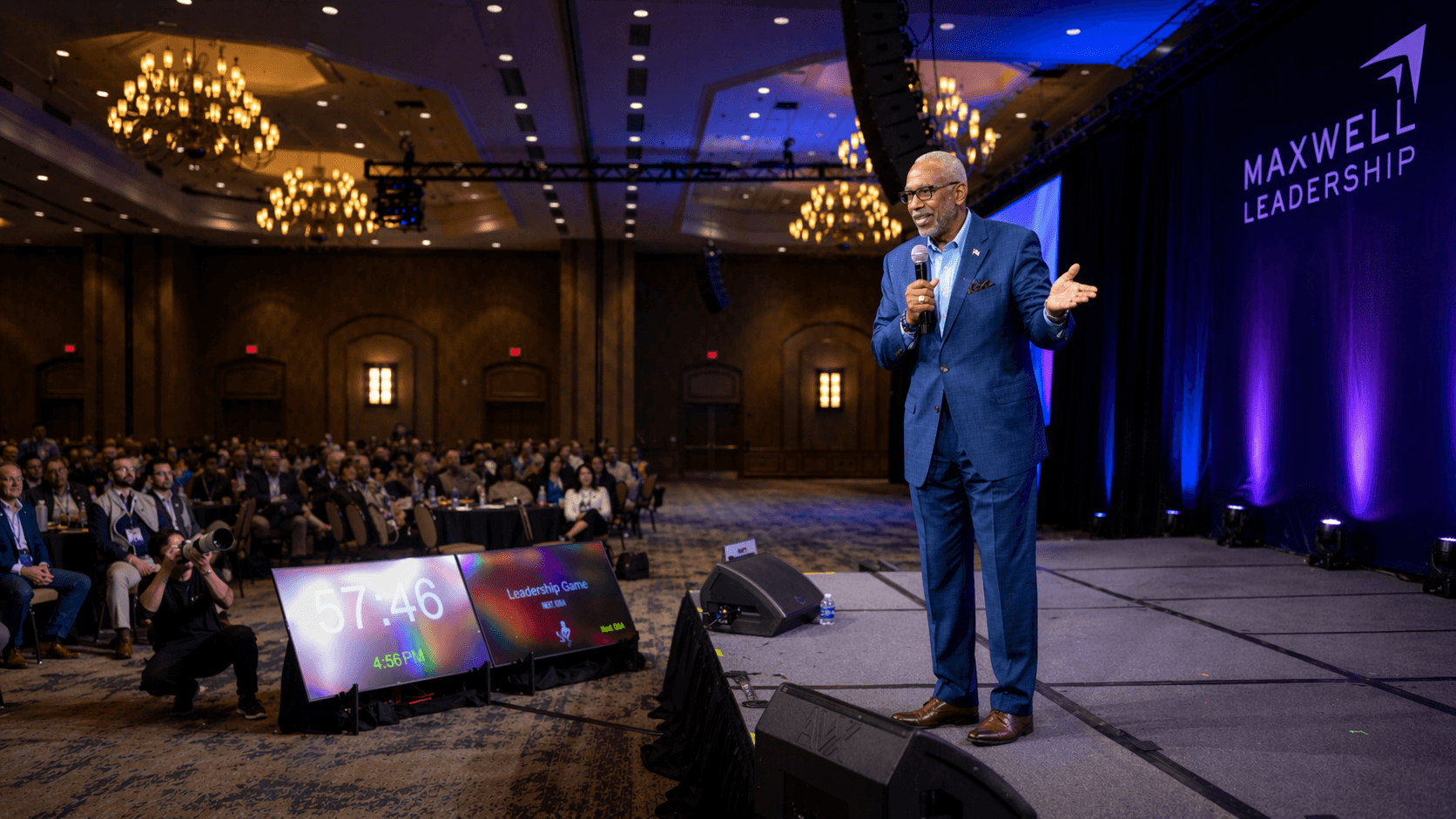 Man in blue suit speaking on stage at conference with audience and MaxWell Leadership sign, dark background, chandeliers.