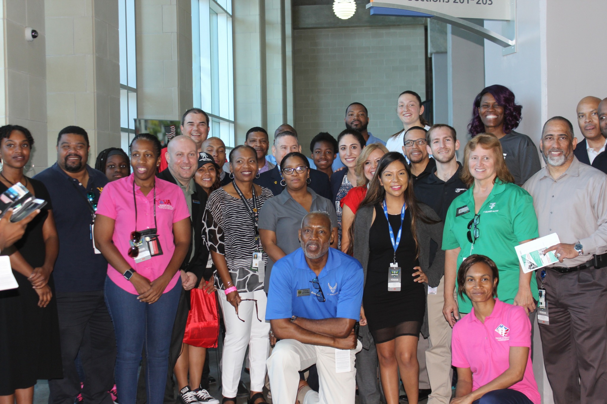 Group of diverse people standing indoors, smiling, some wearing name tags, in a well-lit space with large windows.