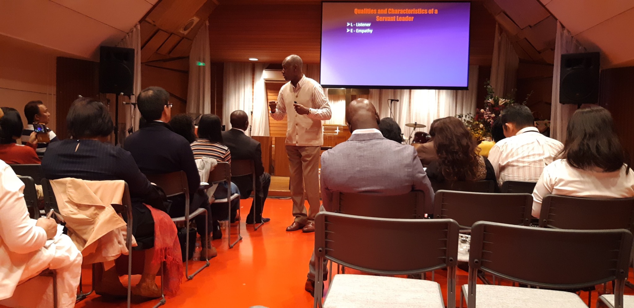 Person standing and speaking to an audience in a room with a purple presentation screen, seated people listening.