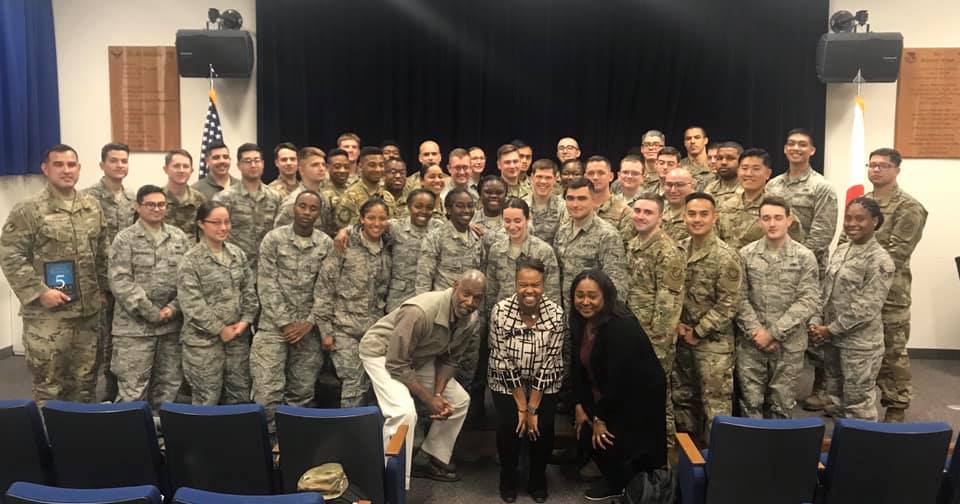 Group of military personnel and civilians posing for a photo in an auditorium with a stage and American flag.