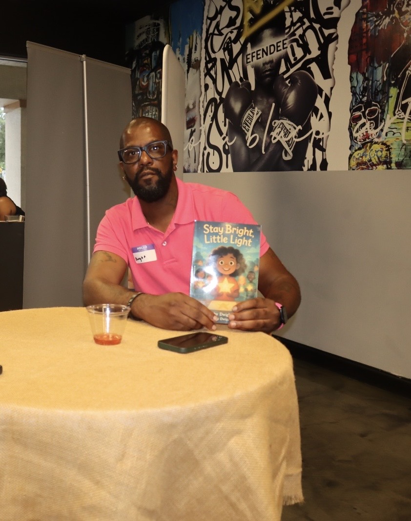 Man with glasses and beard in pink shirt sitting at table holding a children's book, with a drink and phone on table.