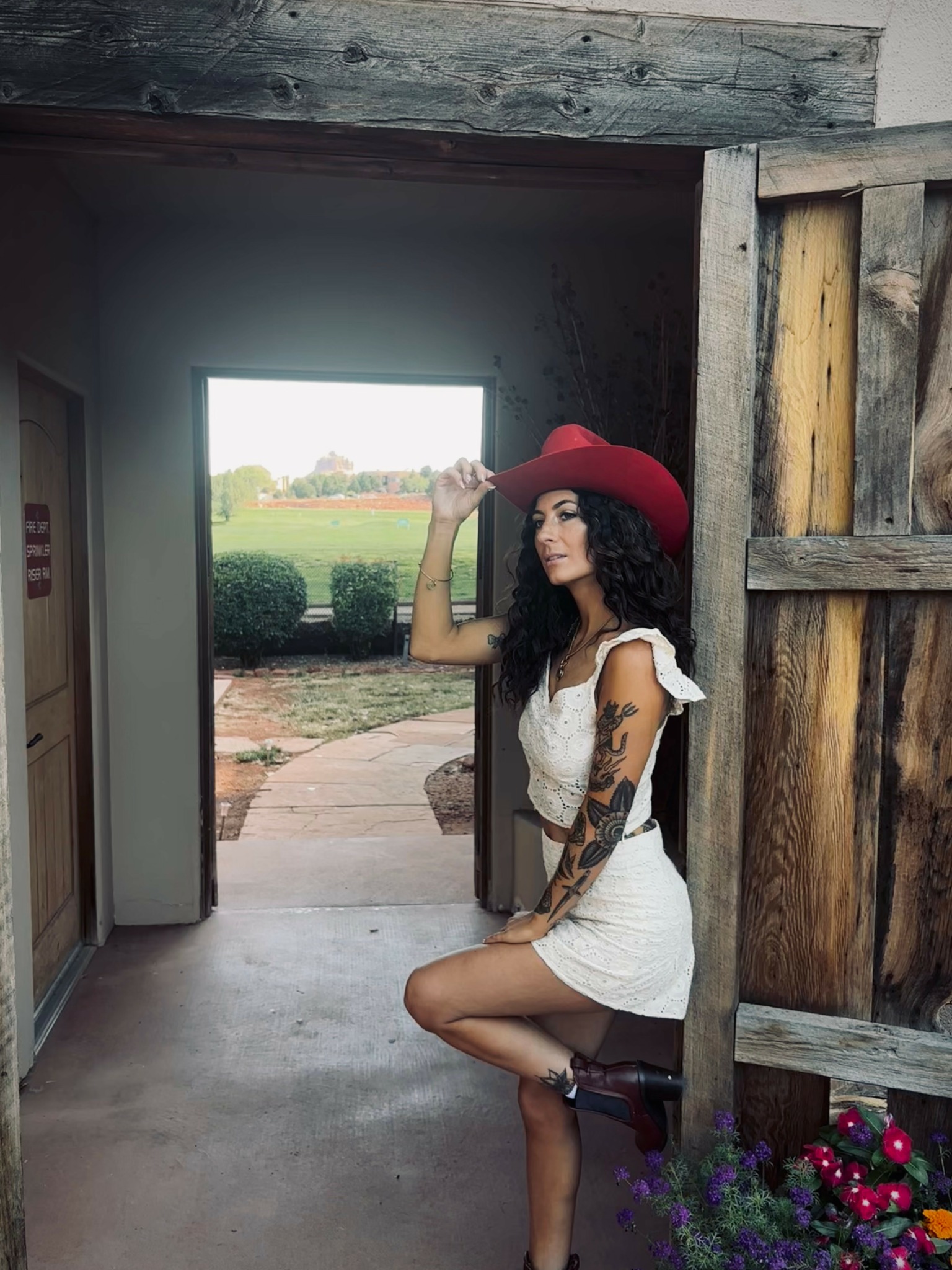 Woman in white dress and red hat standing in doorway, holding hat, with garden view outside, wooden door frame visible.