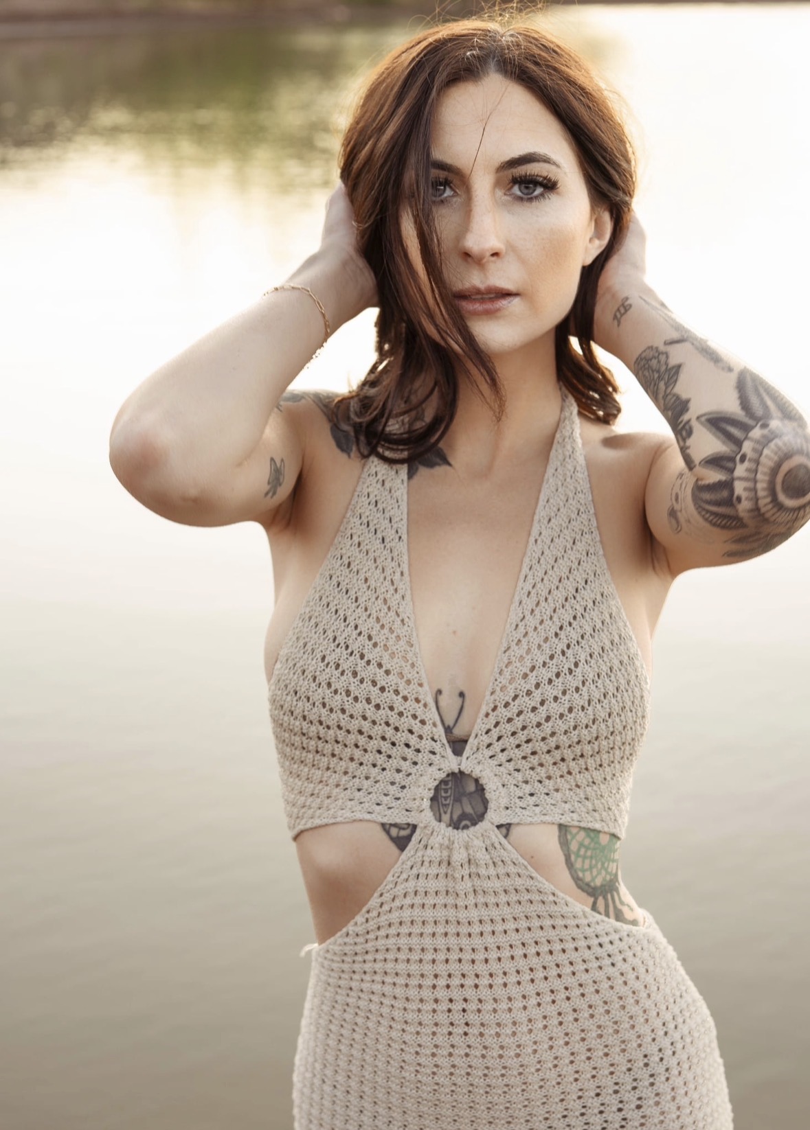Woman with shoulder-length brown hair and tattoos on arms, wearing a beige crochet dress, standing outdoors near water.