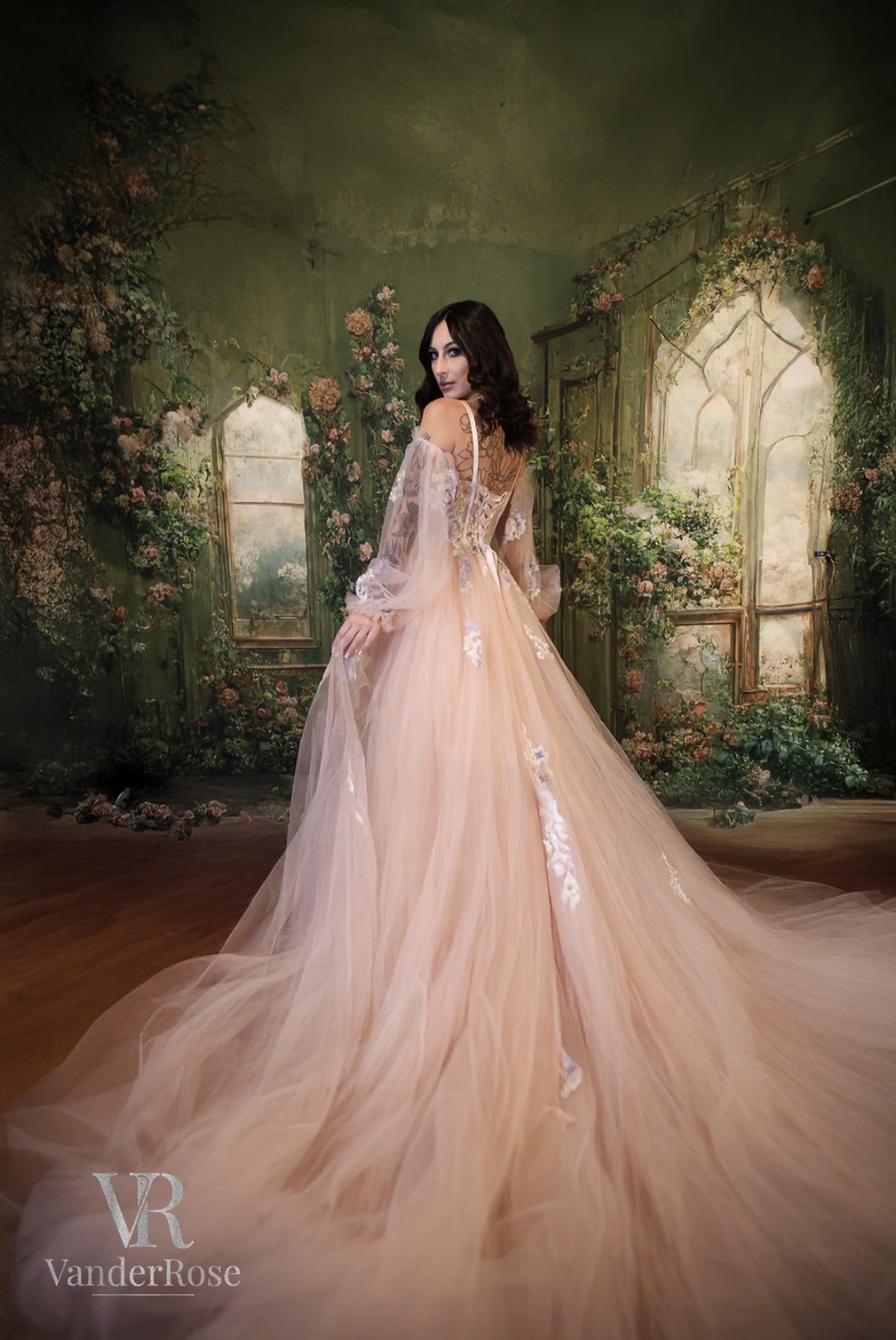 Woman in a flowing, peach-colored gown with lace details, standing in a garden with arched windows and greenery.