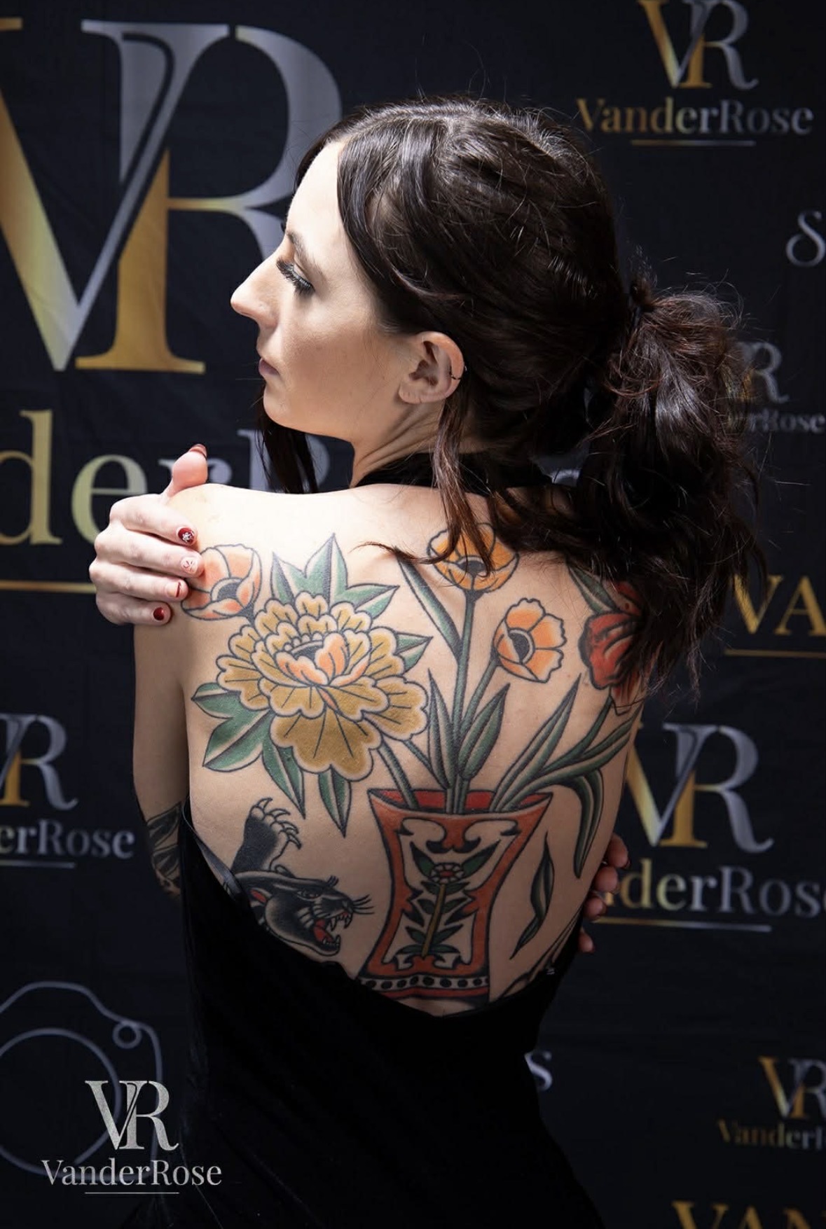 Woman with dark hair and tattoos on her back, facing left, in front of a dark background with logos.