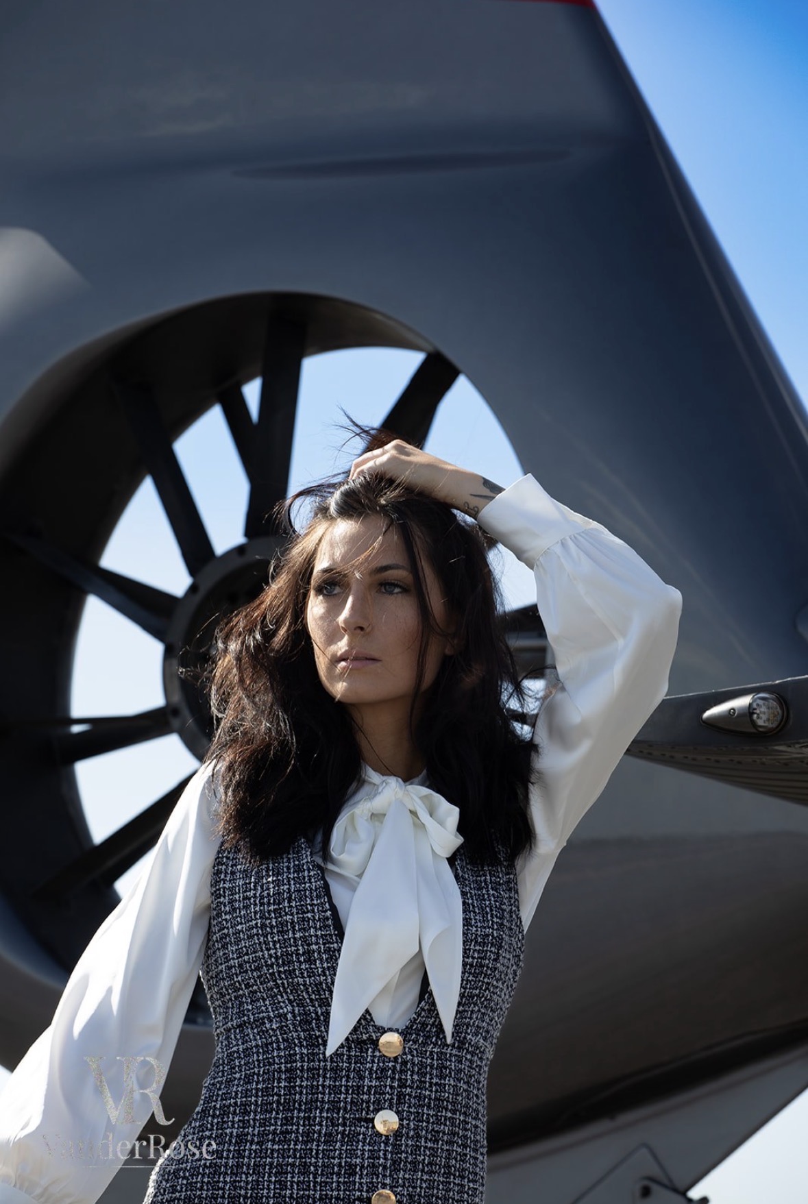Woman with dark wavy hair wearing a white blouse and checkered vest, standing outdoors near a large aircraft wheel, with blue sky in background.
