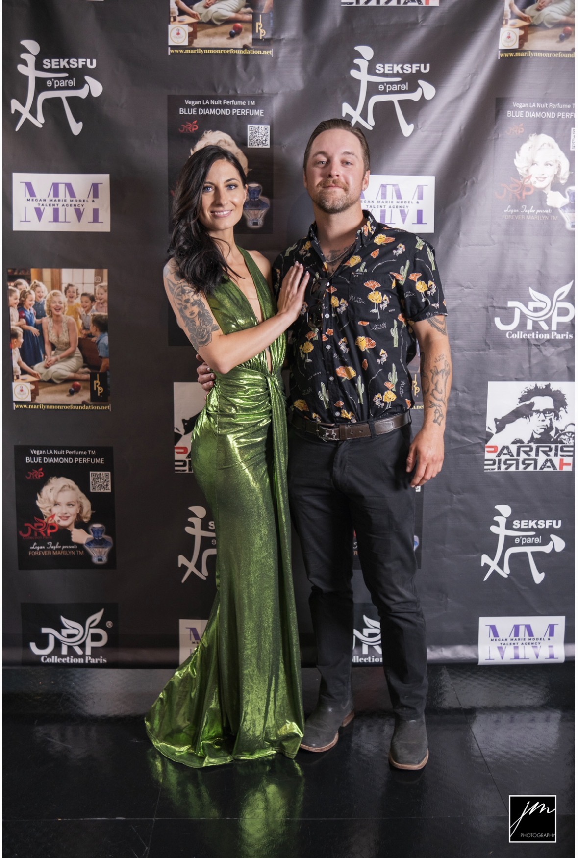 Two people standing in front of a black backdrop with logos, one woman in a green dress and one man in a floral shirt.