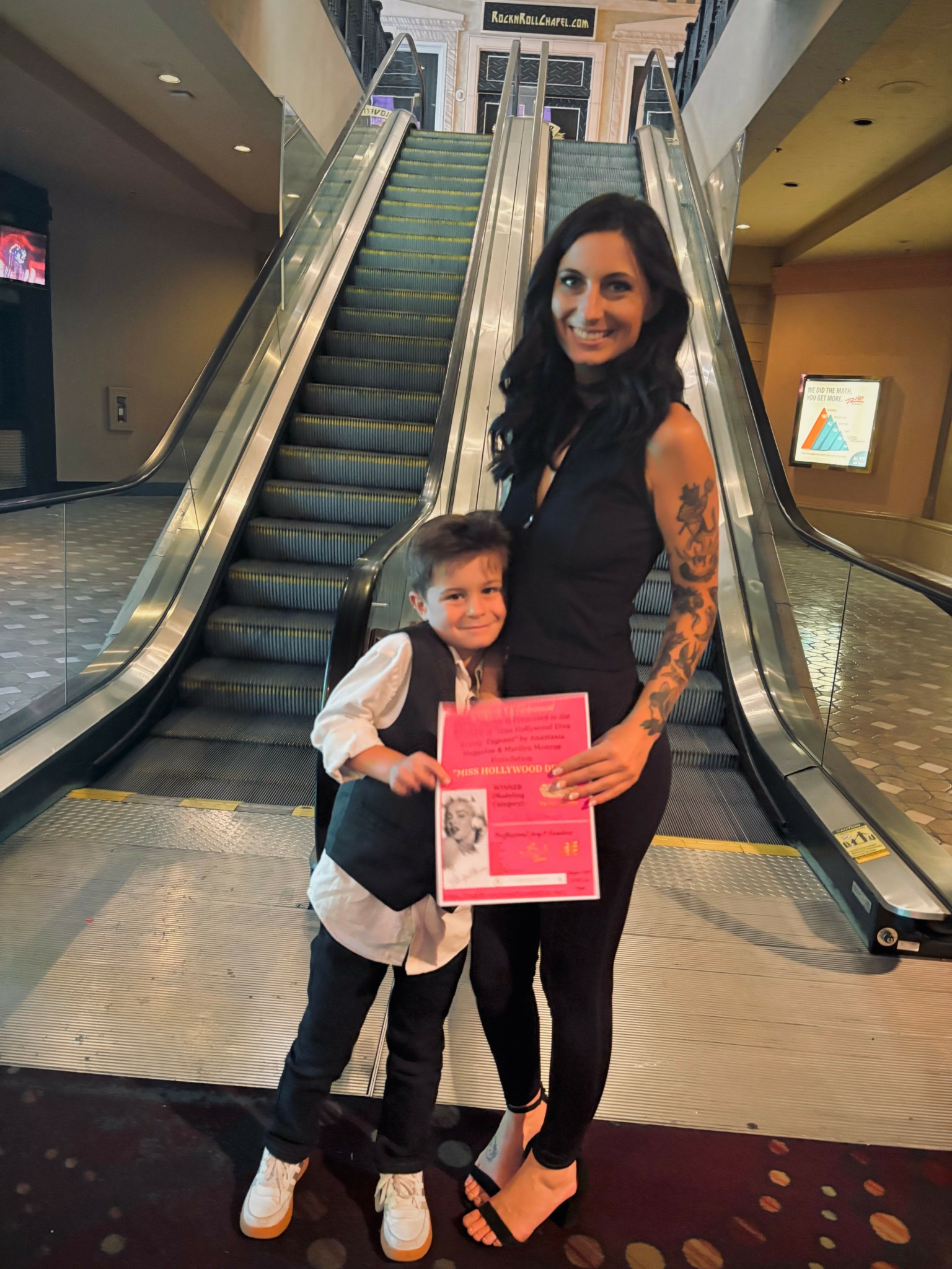 A woman with long dark hair and tattoos on her arm stands on an escalator with a young boy holding a pink certificate, both smiling.
