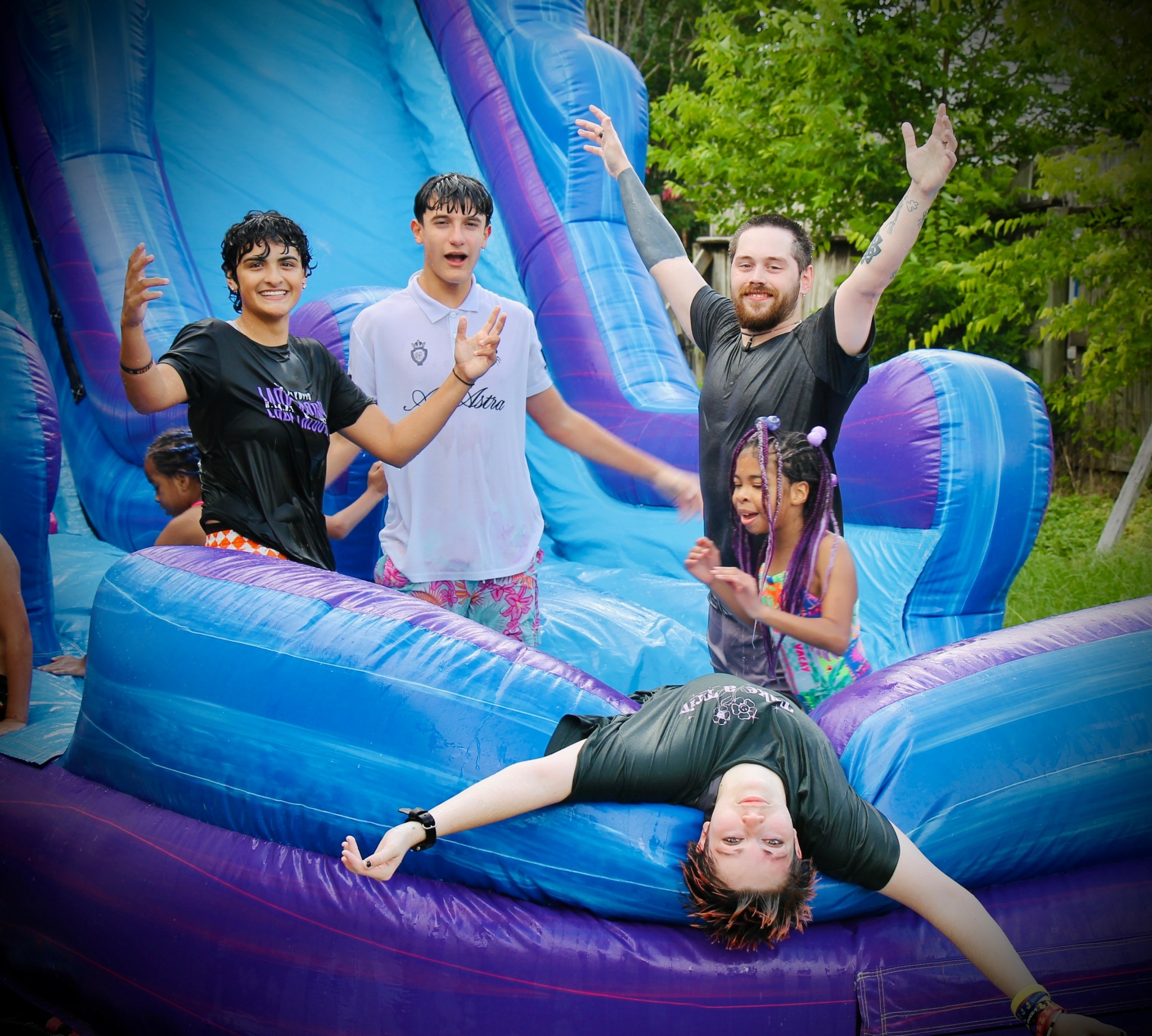 Group of children and an adult on a colorful inflatable slide outdoors, smiling and waving, with trees in background.