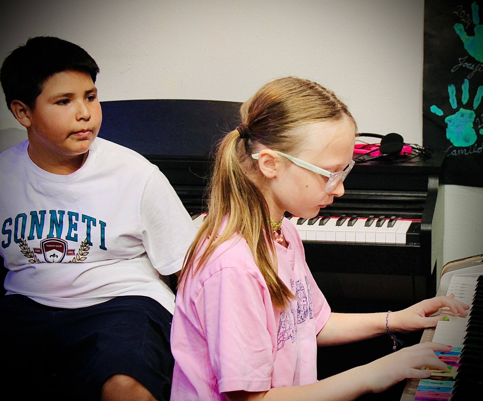Two children, a boy and a girl, sitting near a piano, girl playing the keyboard, boy watching, plain background.