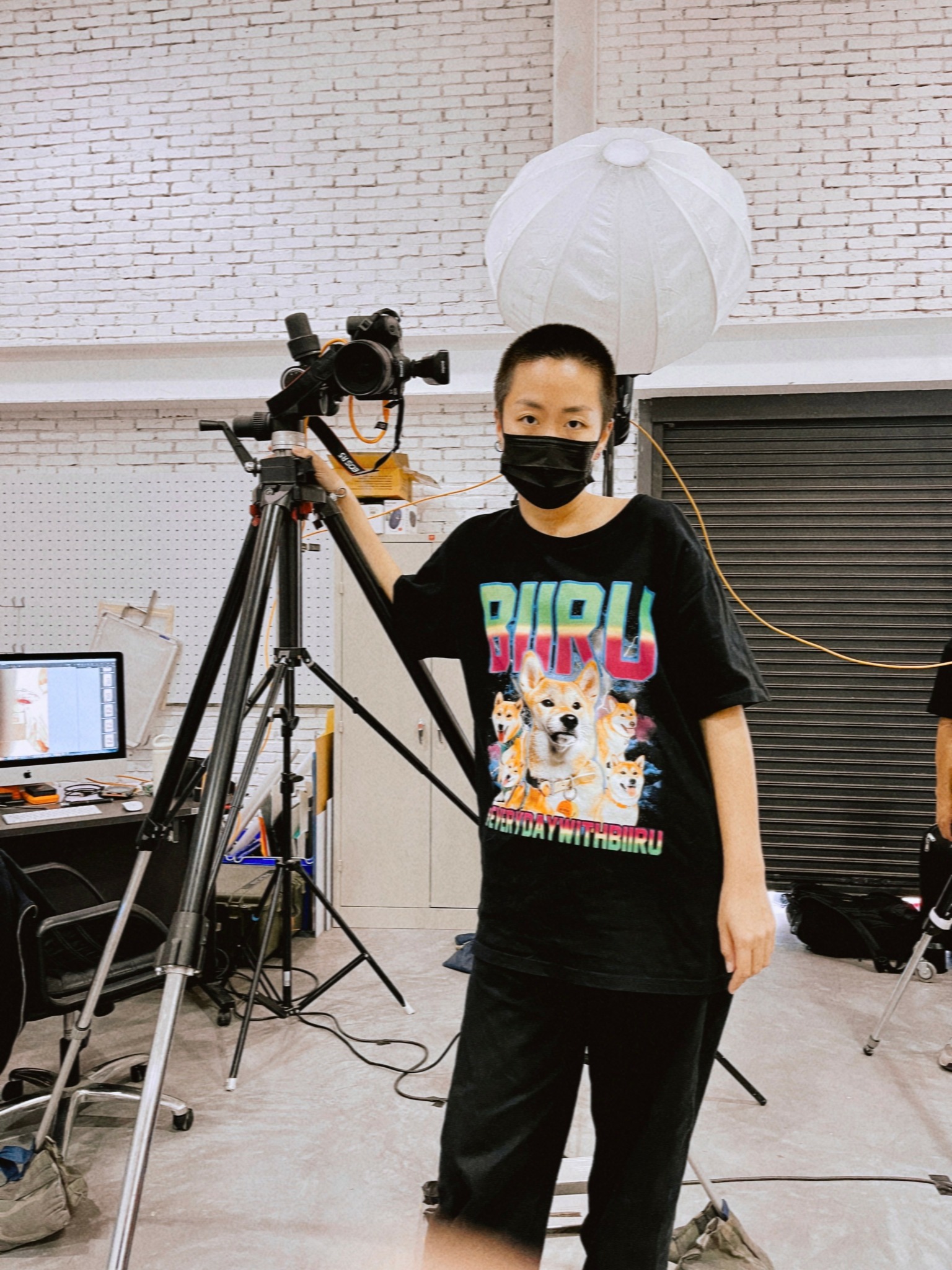 Child wearing a black mask and T-shirt with a dog graphic, standing next to a camera on a tripod in a studio.