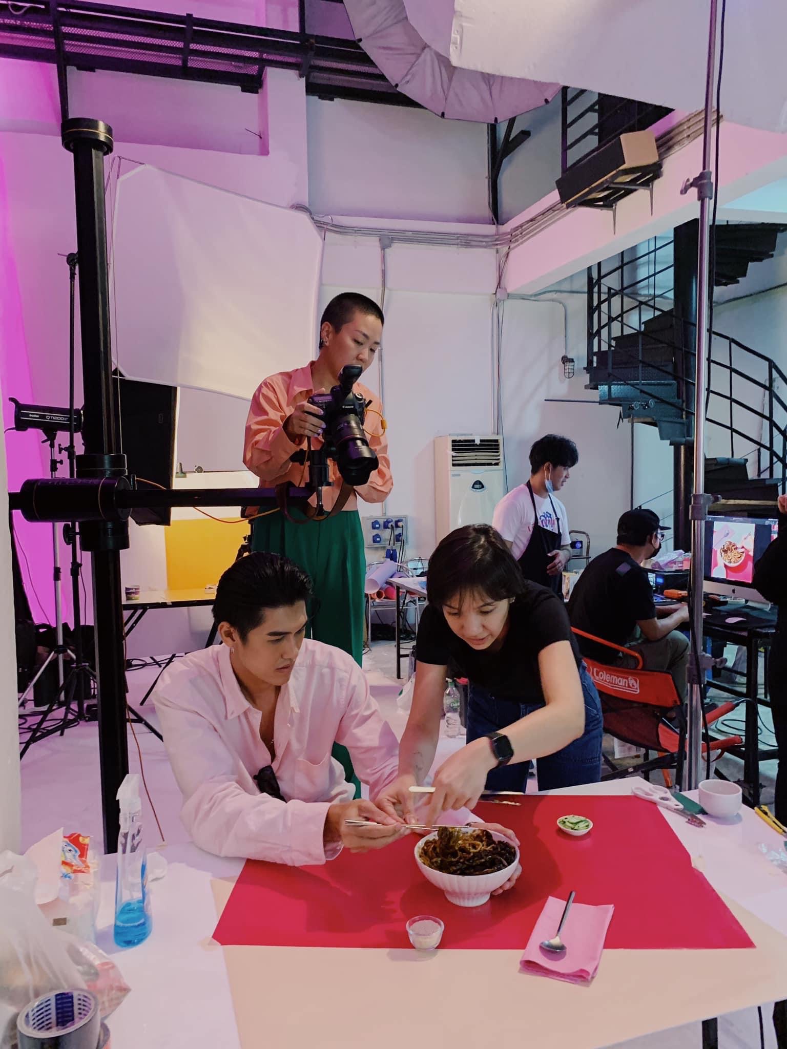 Two people sit at a table with a bowl of food, one in pink and the other in black, with a person filming in the background.
