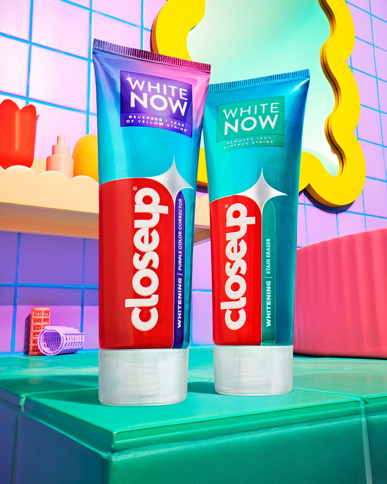 Two tubes of Closeup toothpaste on a green surface with colorful background, including a mirror and shelves with bottles.