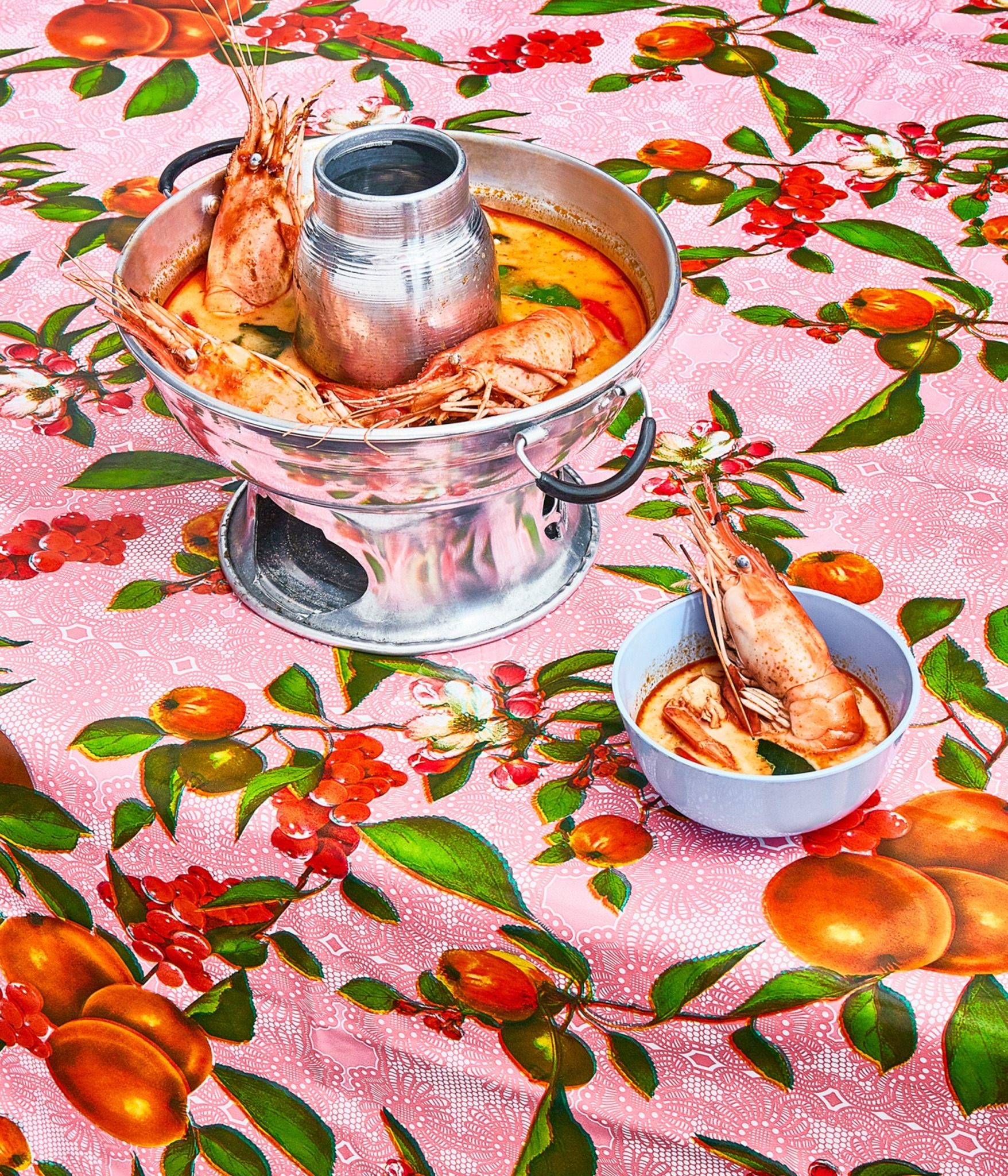 Shrimp dish in a metal pot and a bowl on a floral tablecloth with pink background, orange and green pattern.