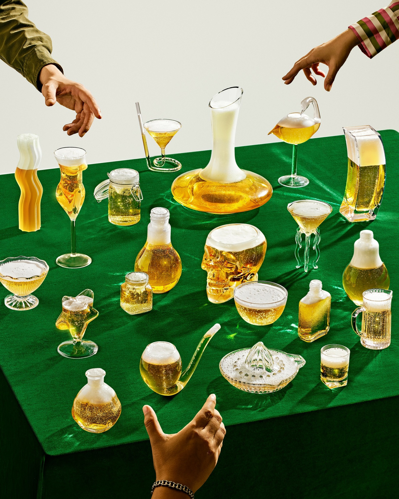 Hands reaching for various drinks and glasses on a green tablecloth, with some glasses filled with beer and others with cocktails.