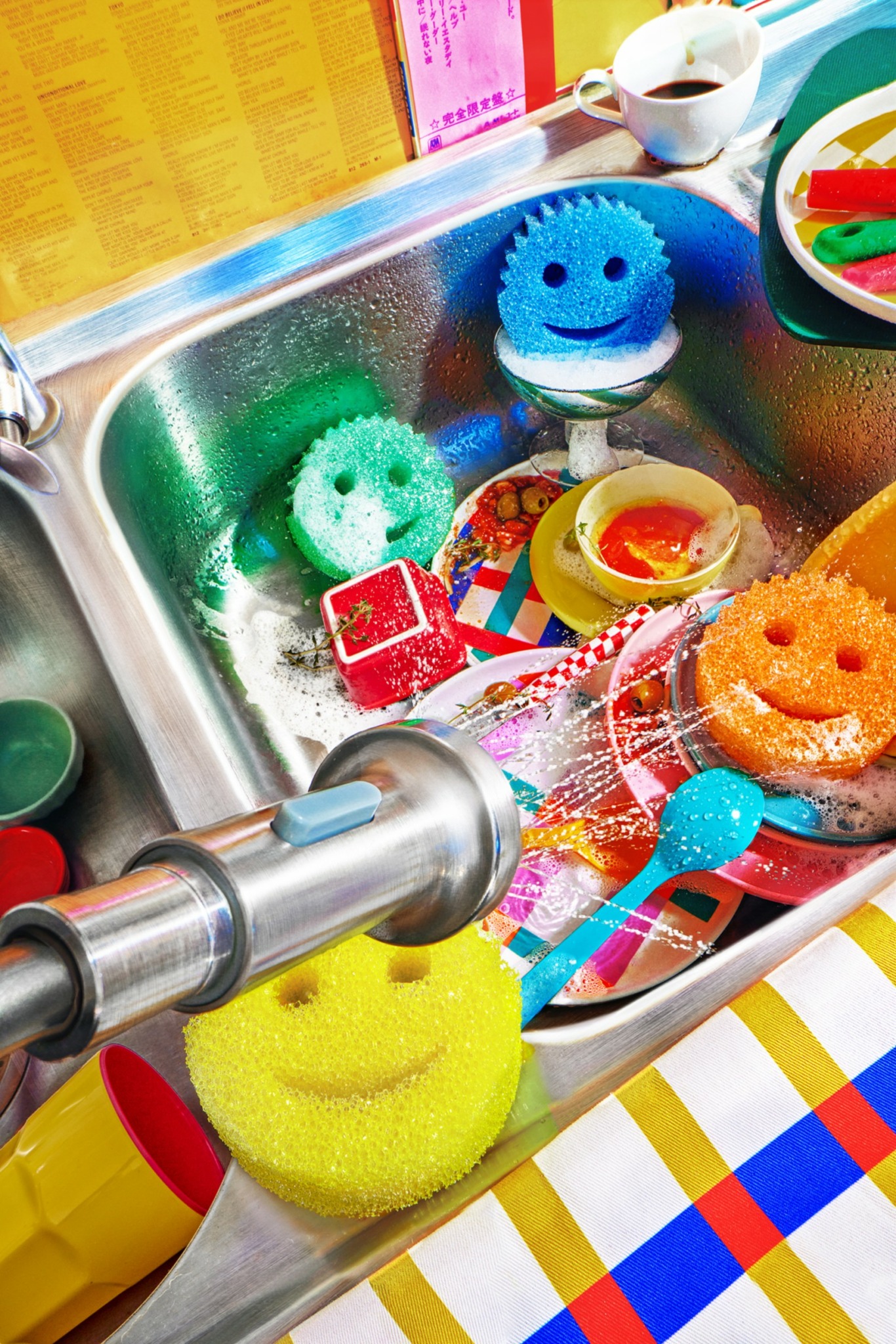 Colorful cleaning sponges with smiley faces in a kitchen sink, surrounded by cleaning supplies and dishes.