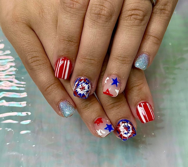 Hands with patriotic-themed nail art featuring red, white, and blue colors, stars, and fireworks designs, on a holographic background.