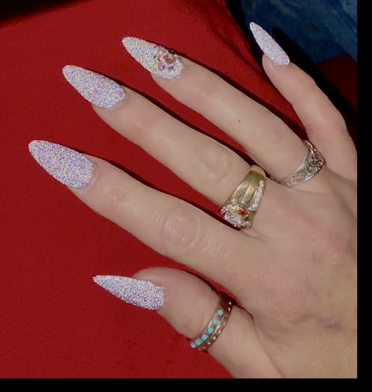 Hand with long, glittery, pointed nails, wearing multiple rings, against a red background.